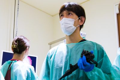 手術着とマスクを着用した医師が、内視鏡を手に持ち、モニターを真剣な表情で見上げながら検査を行っている。精密な医療技術と専門性の高さを感じさせる手術室での一場面。