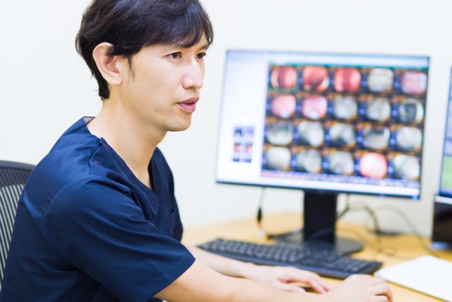 スクラブを着た男性医師が、デスクでPCモニターに表示された内視鏡の画像を確認しながら、診断結果を説明している。先進的な医療技術を用いた精密な検査と、専門医による的確な診断の重要性を示す場面。
