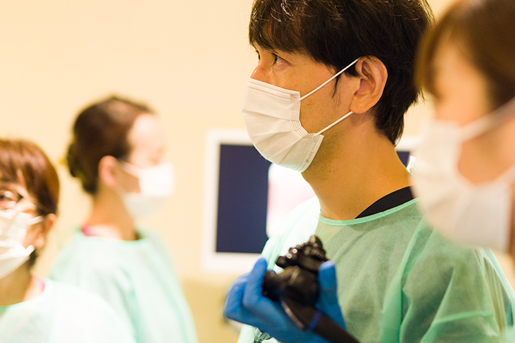 手術着とマスクを着用した医師が、内視鏡を手に持ち、真剣な眼差しで前方のモニターを見つめている。周囲の医療スタッフと共に、チーム医療で高度な検査に臨む緊張感と専門性が伝わる手術室の光景。