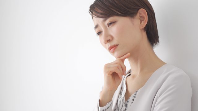 短髪の日本の女性が、あごに手を当てて白い壁の前で考え込んでいる。