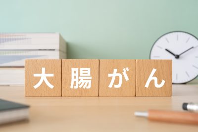 木製のブロックが机の上に置かれており、「大腸がん」という漢字とひらがなの文字が書かれている。背景には時計、本、その他の学習用品が写っている。